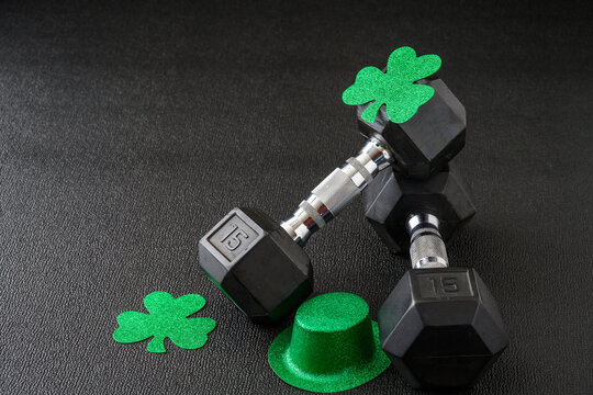 Set of hex head dumbbells with green glitter shamrocks and leprechaun hat on a black gym floor, happy St. Patrick’s Day
