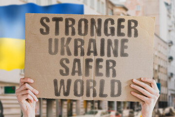 The phrase " Stronger Ukraine, Safer World " is on a banner in men's hands with blurred background. Support. Encouragement. Ukrainian. Strength. Alliance. International. Policy. Strategy. Freedom