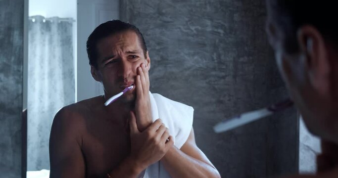 Young man looking in mirror brushing teeth in bathroom and feel dental pain discomfort suffer from toothache. Concept of caries problem with teeth oral disease unwell illnesses. Dental problems