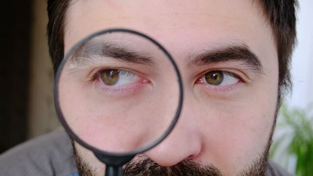 Close up portrait of attractive man looking through magnifying glass at camera . Male searching sales or discounts. Shopping and detective concept. High quality FullHD footage with slow motion. 