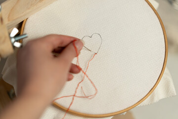Manos bordando un corazón en tela cuadrillé blanca en bastidor de madera redondo con hilo rojo 