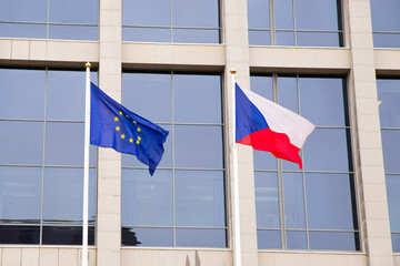EU flag and Czech Republic flag on flagpole on modern consulate building. Flag of European Union and Czech flag on glass blue facade. - obrazy, fototapety, plakaty