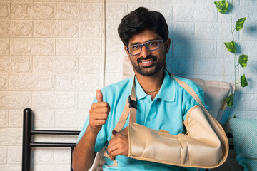 happy smiling young man with broken hand at bedroom showing thumbs up gesture by looming at camera...