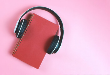 flat lay of red notebook or  diary or planner covered with headphones on pink background with copy space.  Audio book or podcast concept.