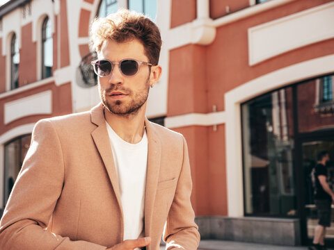 Handsome confident stylish hipster model.Sexy modern man dressed in elegant beige suit. Fashion male posing in the street background in Europe city at sunset. In sunglasses