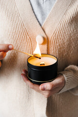 Soy scented candles in metal jars, boxes. Candle held by a girl against a sweater. A long match burns.  Modern handmade coconut wax candles without paraffin. 