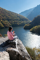 Young woman sitting alone on edge of cliff relaxing enjoying amazing scenic view of mountain river valley. Rhodopes mountains, Bulgaria. Weekend nature getaway, travel, vacation outdoor, hiking