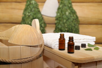 Bottles of healing essential oil stand on a wooden bench next to a towel against the background of sauna accessories hanging on a log wall. The concept of natural aromatherapy, sauna and spa treatment
