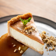 Cheesecake with caramel. Piece of dessert sprinkled with nuts and decorated with mint on plate. Sweets with salted caramel. Wooden table. Side view. Soft focus. Copy space. High quality photo