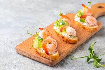 Open sandwiches on white bread with cream cheese, fresh cucumber, shrimp and green pea microgreens on a wooden board on a gray concrete background.