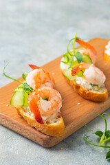 Open sandwiches on white bread with cream cheese, fresh cucumber, shrimp and green pea microgreens on a wooden board on a gray concrete background.