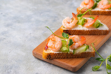 Open sandwiches on white bread with cream cheese, fresh cucumber, shrimp and green pea microgreens on a wooden board on a gray concrete background.