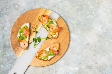 Open sandwiches on white bread with cream cheese, fresh cucumber, shrimp and green pea microgreens on a wooden round board on a gray concrete background. Scandinavian cuisine