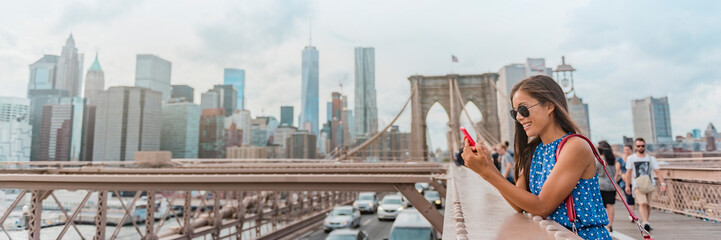New York woman using phone app walking on Brooklyn Bridge by Manhattan city skyline. Young female professional multicultural lady, New York City, USA. Panoramic banner - obrazy, fototapety, plakaty