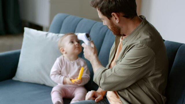 Young caring father measuring body temperature of his playful baby daughter and seeing fever, empty space