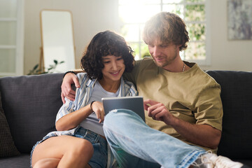 Young affectionate couple in casualwear scrolling through online goods or videos in digital tablet while sitting on couch in living room