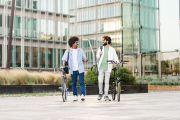 Two young adults walking together on the way to work. Young healthy friends with bikes going to the office  - obrazy, fototapety, plakaty