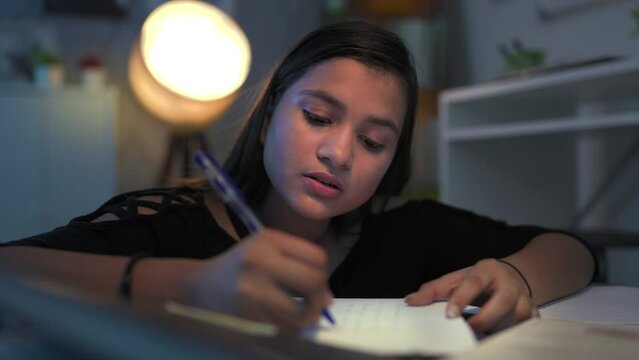 Young Indian college girl with pen in hand writing, studying, thinking, doing assignment worried for term exam. Troubled teenager revise lesson late hours in the night at home that doesn&rsquo;t know answer