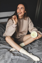 A young woman lies on her bed while eating a pint of pistachio ice cream with spoon. She is lick spoon with ice cream. Eating in bed. Happy beautiful woman resting in her comfortable bed at home. 