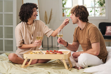 Happy young woman with flute of champagne putting piece of snack in mouth of her husband while both enjoying breakfast in bed
