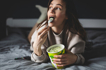 A young woman lies on her bed while eating a pint of pistachio ice cream with spoon. She is lick spoon with ice cream. Eating in bed. Happy beautiful woman resting in her comfortable bed at home.  - obrazy, fototapety, plakaty