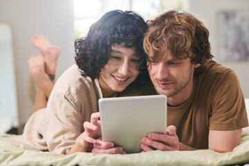 Young affectionate couple looking through online goods on tablet screen while relaxing on bed after waking up in the morning on weekend