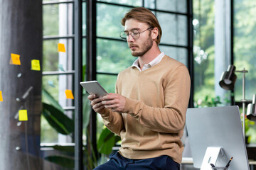 Fototapeta premium Young successful thinking programmer inside loft style office uses tablet computer, young blond man in casual clothes reads news online, serious businessman browses internet pages.