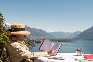 Woman with Straw Hat Sitting and Reading the Menu in a Restaurant with Panoramic View over Lake Maggiore in a Sunny Day in Ascona, Ticino, Switzerland.
