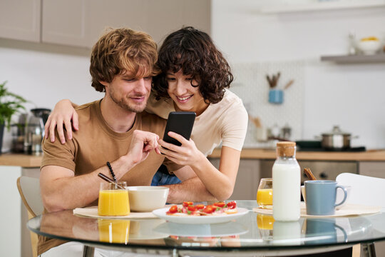 Young smiling woman showing something in smartphone to her husband sitting by table with sandwiches, juice and milk and having breakfast