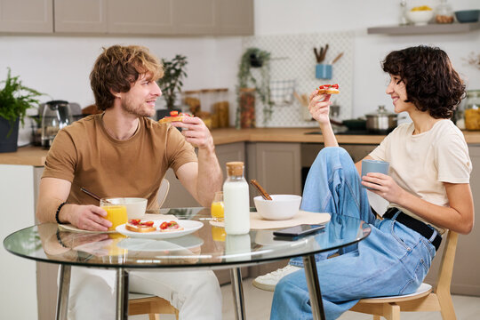 Happy young couple having vegetable sandwiches and juice for breakfast while sitting by table in the kitchen and discussing plans for the day