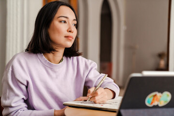 Naklejka premium Pensive asian woman artist drawing in sketchbook while sitting at table at home