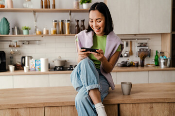 Excited asian woman playing online game on mobile phone while sitting in kitchen