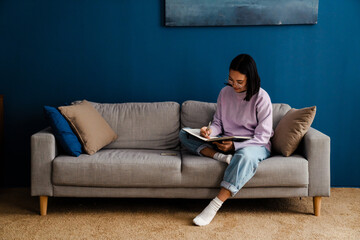 Asian woman writing down notes while sitting on sofa