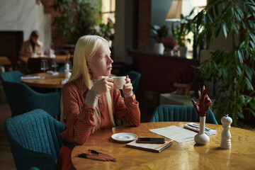 Young pensive albino woman with cup of tea sitting in comfortable armchair by table in cozy cafe decorated with green plants