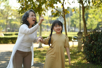 Joyful little girl and grandmother walking in public park surrounded by green trees at sunlight morning. Family, generation concept