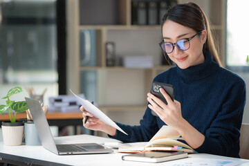 Beautiful Asian woman using a smartphone and working with a laptop while sitting at an office desk, working from home concept.