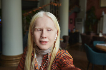 Portrait of young albino woman with earphones looking at camera while holding mobile phone in front of her face and talking in video chat