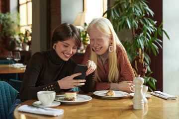 Two young cheerful girlfriends discussing online photo or video in mobile phone while sitting by table in cozy cafe and having dessert with tea