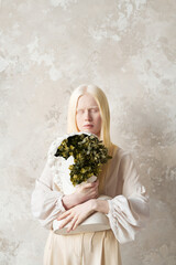 Young serene albino woman in white attire holding plastic bust with green plants inside while standing against wall with stylish wallpapers