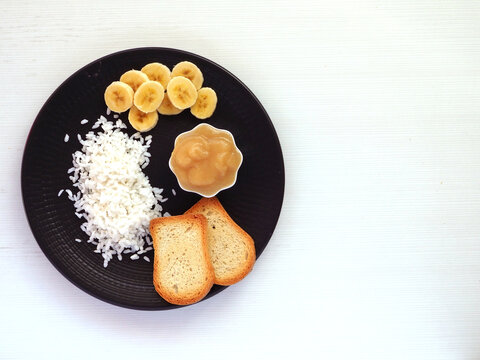 Banana slices, boiled rice, applesauce and toast on a dark plate. Food for BRAT diet.