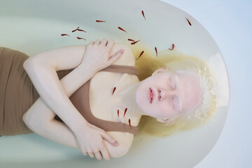 Above view of young pale woman with albinism lying in clear water in bathtub while stock of tiny...