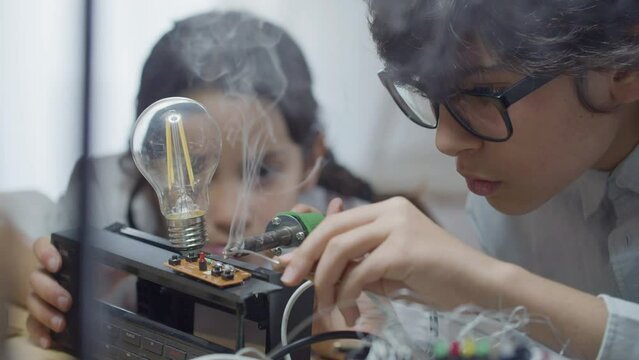 Two smart kids practicing their hobby in electrical engineering. Boy in glasses accurately soldering circuit board with tin solder wire while girl watching process. Hobby, smart children concept 