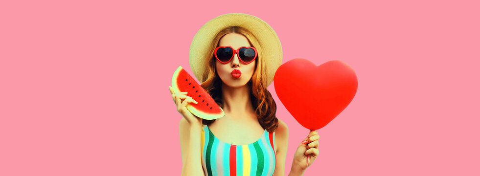 Portrait of beautiful young woman with slice of fresh watermelon and big red heart shaped balloon blowing her lips wearing summer straw hat, sunglasses on pink background