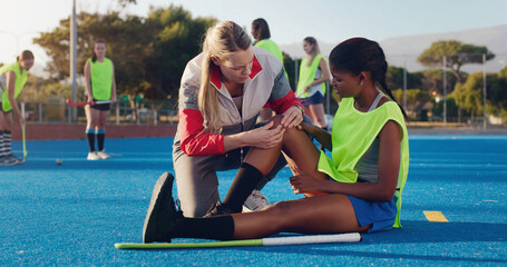 Woman, sports and knee injury in hockey training, practice or game on a blue turf with coach and team. Sport mentor helping female in leg pain, accident or bruise from physical activity during match - Powered by Adobe