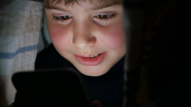 A caucasoid teenager guy at a late hour, covered with a blanket, is talking via video link with a friend using a smartphone. The influence of gadgets on vision and nervous irritability and depression