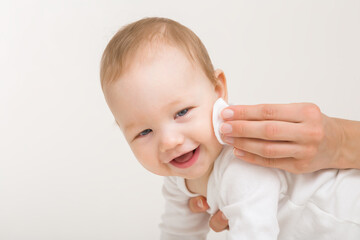 Young adult mother hand wiping baby boy cheek with white cotton pad. Light gray background. Daily...