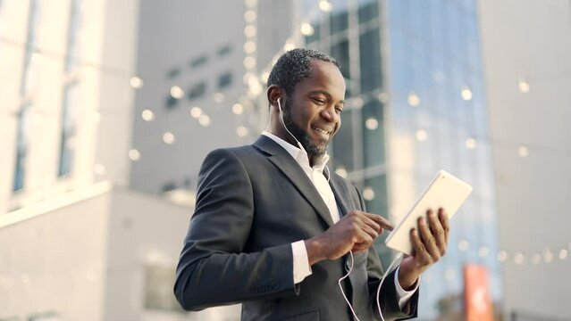 Smiling african american businessman with headphones using tablet while standing outside. Mature confident businessman in formal suit reading good news while texting online in front of office building