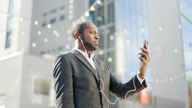 Serious african american businessman with headphones communicates online using smartphone while standing outside. A mature confident male in a formal suit is negotiating in front of an office building