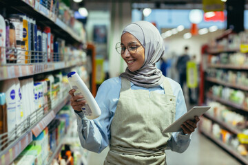 A young Arab woman in a hijab works in a supermarket, salesperson, consultant, standing with a...