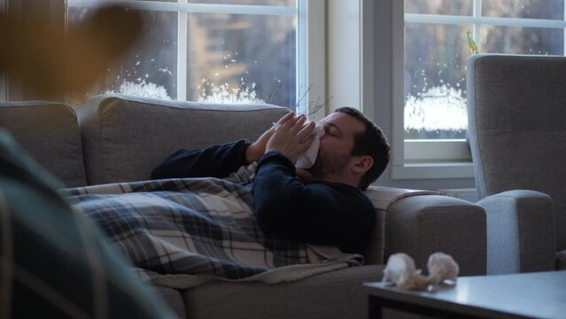 Man with flu sneezing into napkin while lying down on sofa in living room